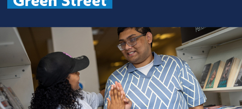 The image shows two people interacting in a library. The person on the left is wearing a black cap and a light gray sweater, while the person on the right is wearing a blue and white patterned shirt. The text at the top of the image reads "People Powered Green Street" and there is also a logo for "Newham London" in the top right corner.