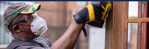 A person wearing a cap, safety glasses, face mask, and gloves is using a power tool to work on a metal structure. The background shows an outdoor setting with blurred buildings. The top part of the image has text that reads "People Powered Forest Gate" in white letters on blue and teal backgrounds. There is also a logo for Newham London in the top right corner.